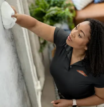 A SERVPRO technician in a black polo shirt wiping down a wall with a white cloth during a professional cleaning service, with indoor plants visible in the background.