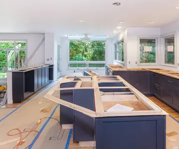 A kitchen under renovation with navy blue cabinets being installed, showing an unfinished island in the foreground, upper cabinet spaces along the walls, and large windows overlooking green trees in the background.