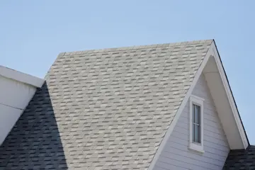 The peaked roofline of a white residential home featuring light gray asphalt shingles, a small gable window, and white siding against a clear blue sky.